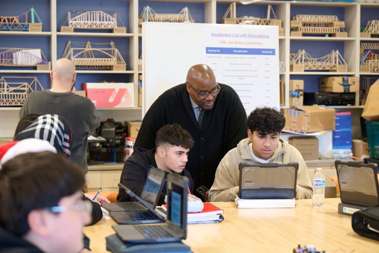 A man looking over the shoulders of students working on computers at a table