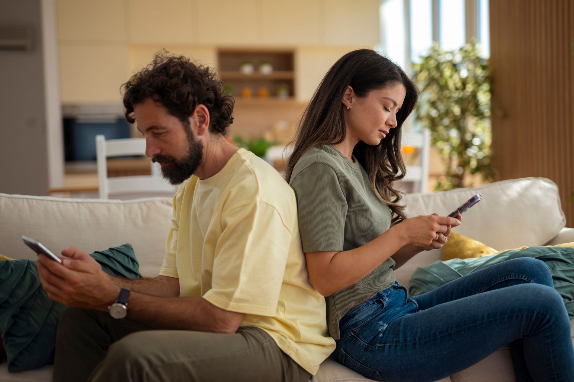 Young couple sitting back-to-back on a sofa and using their phones.