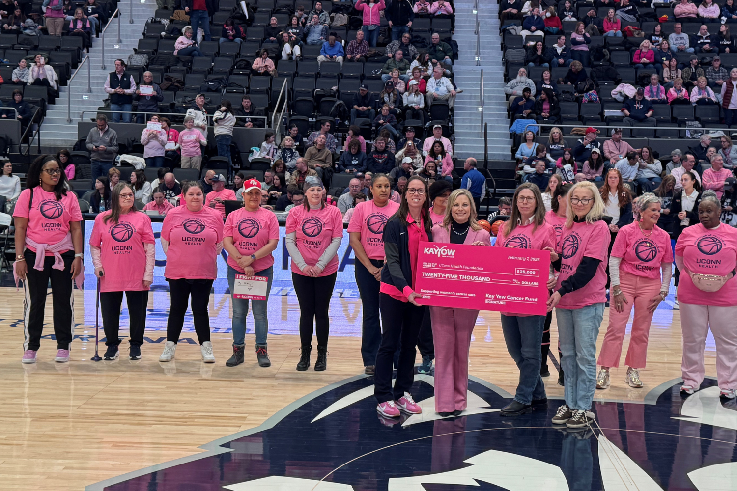 group of women in pink shirts stand on a basketball court holding a large pink check.