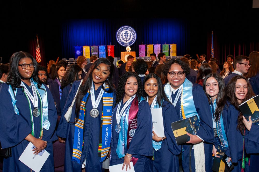 Students in graduation regalia and medals posing for a photo at the front of an auditorium while UConn's president can be seen speaking in the background