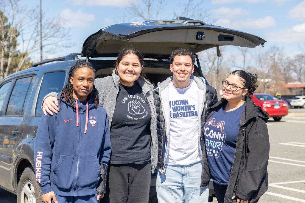 Four young people in UConn apparel pose together in front of a car with an open tailgate.