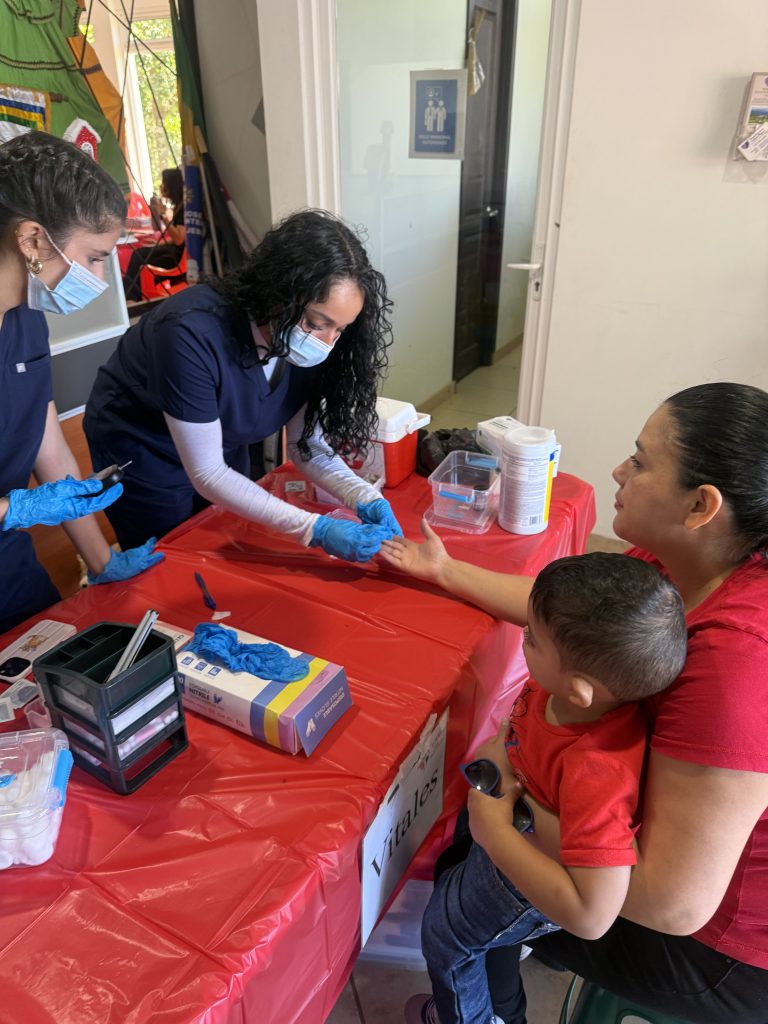 A student in medical mask and rubber gloves swabs the hand of a woman in a medical clinic holding a baby.