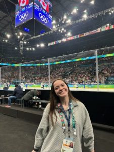 A young woman standing in front of an ice hockey arena with the Olympic rings on the scoreboard above her.