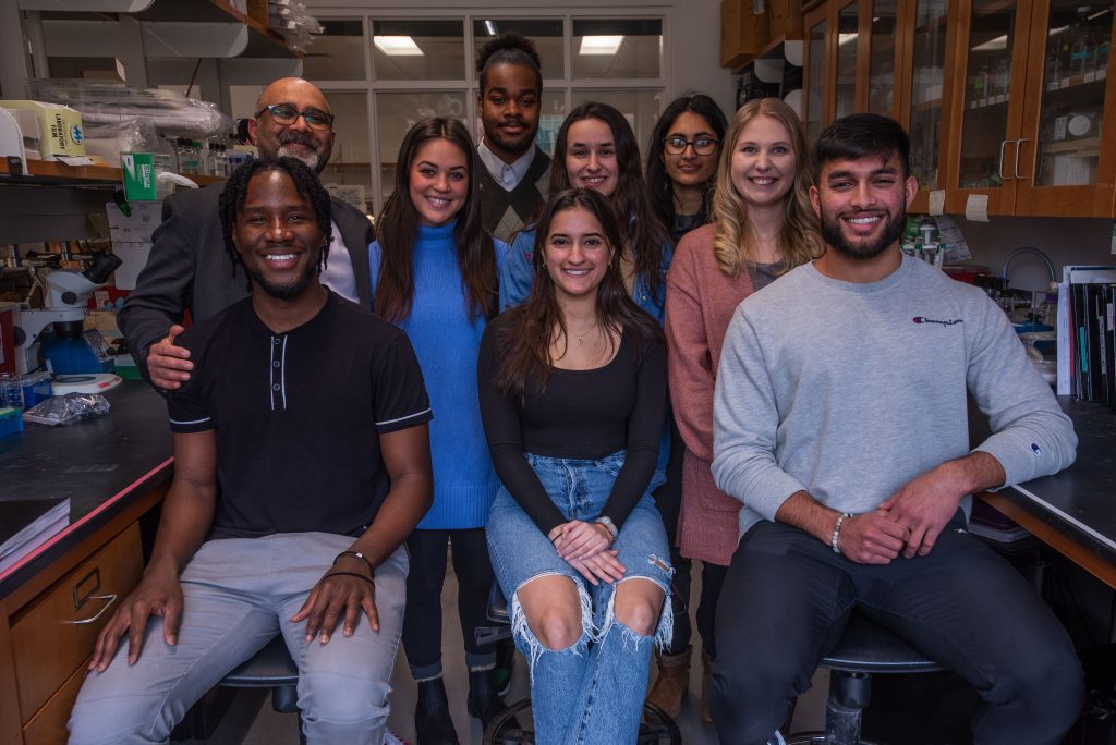 A group of students sitting and smiling in a lab, with Kanadia smiling behind them