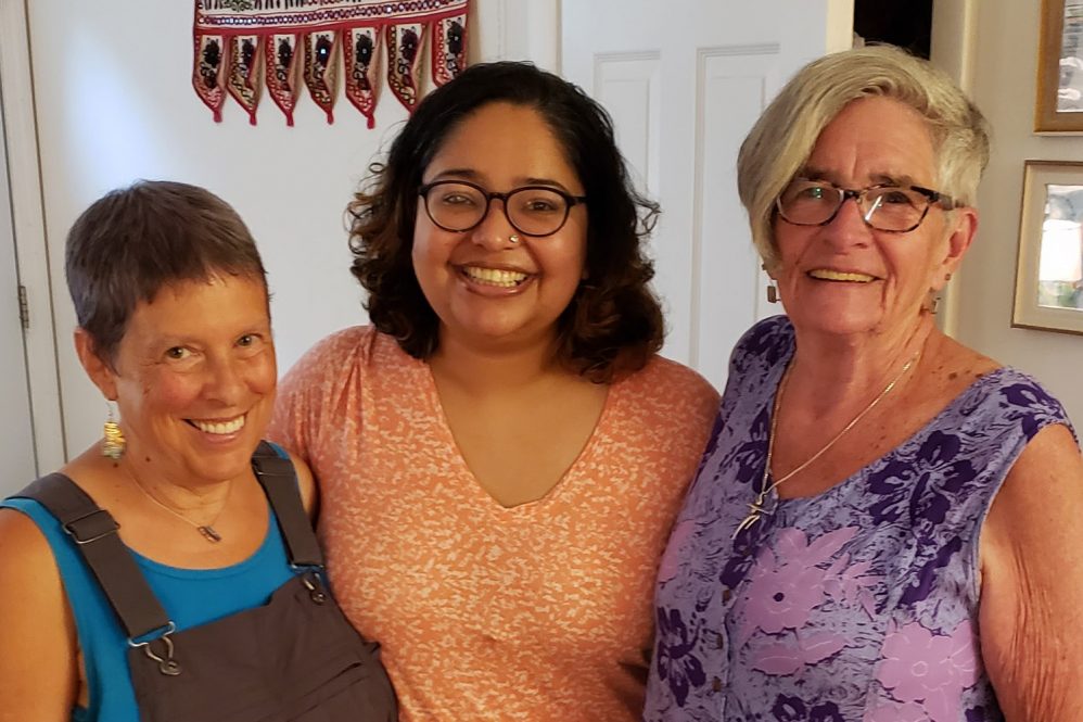 Three people - one in overalls and a blue shirt, another in an orange shirt, and the third in a purple dress, pose arm-in-arm for a photograph.