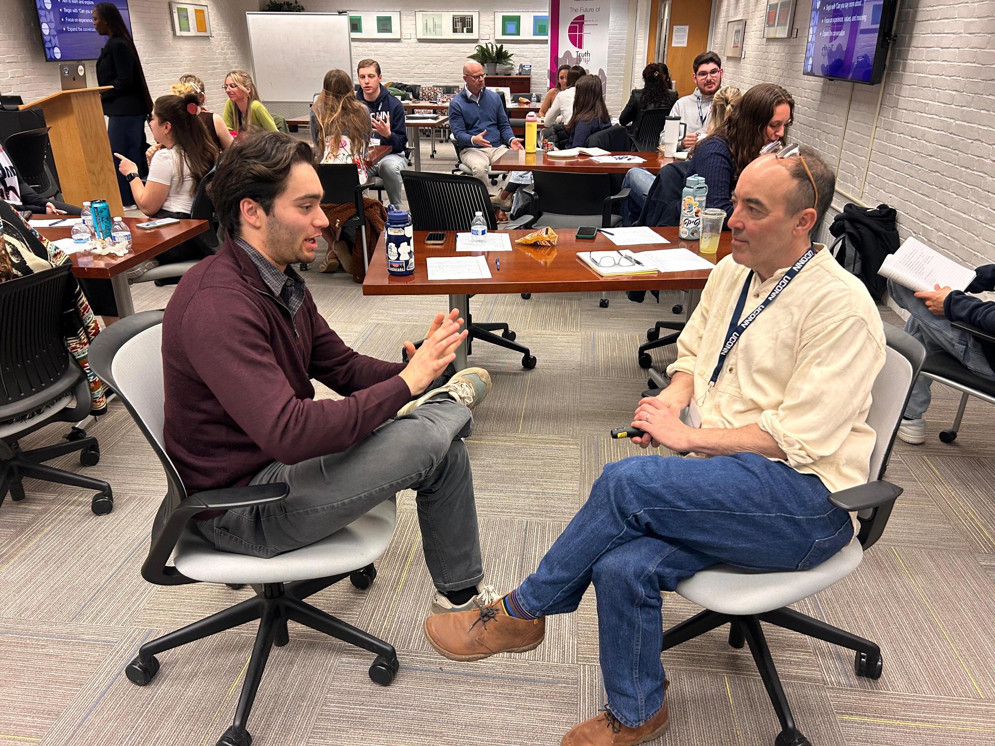 Two men in the foreground sit facing each other in discussion, with a room full of others behind them.