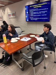 Two people at a table look to their left, to an off-camera speaker.