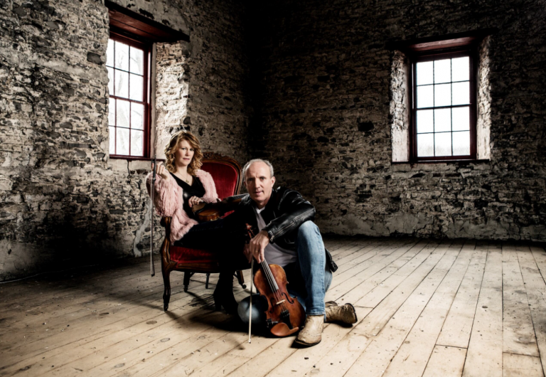 A woman sitting in a chair and a man sitting on the floor in front of her both hold violins.