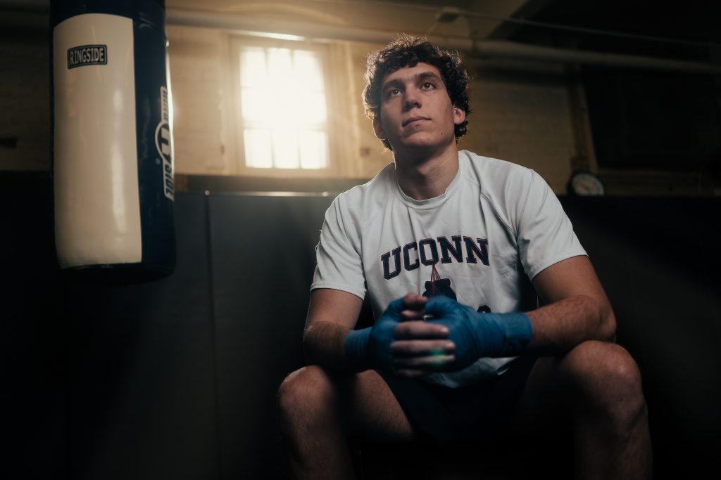 A young man in a UConn t-shirt sits next to a heavy punching bag.