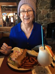 A woman wearing a hat sitting at a table with a lobster roll in front of her 