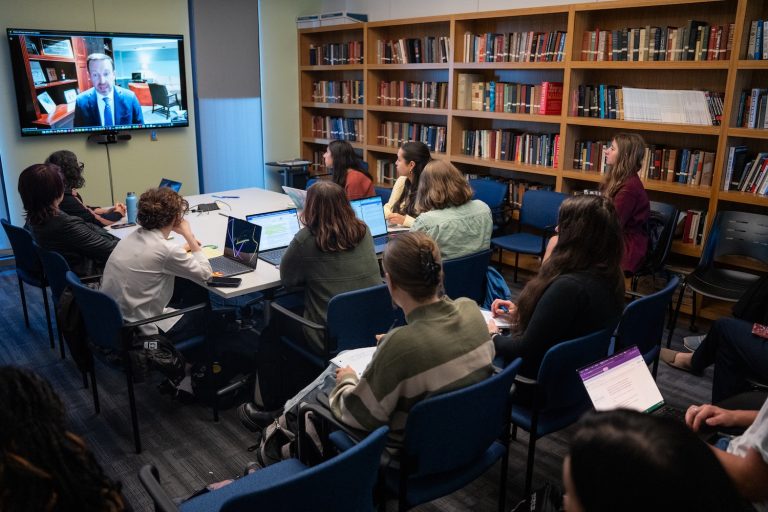 A classroom full of students looks at a television screen depicting a man in a shirt and tie.