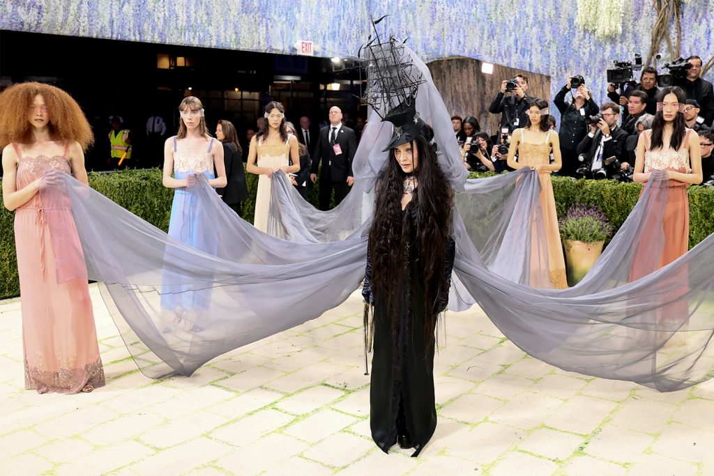 A woman dressed in a black dress and hat with scarves flowing behind her with six women holding the ends
