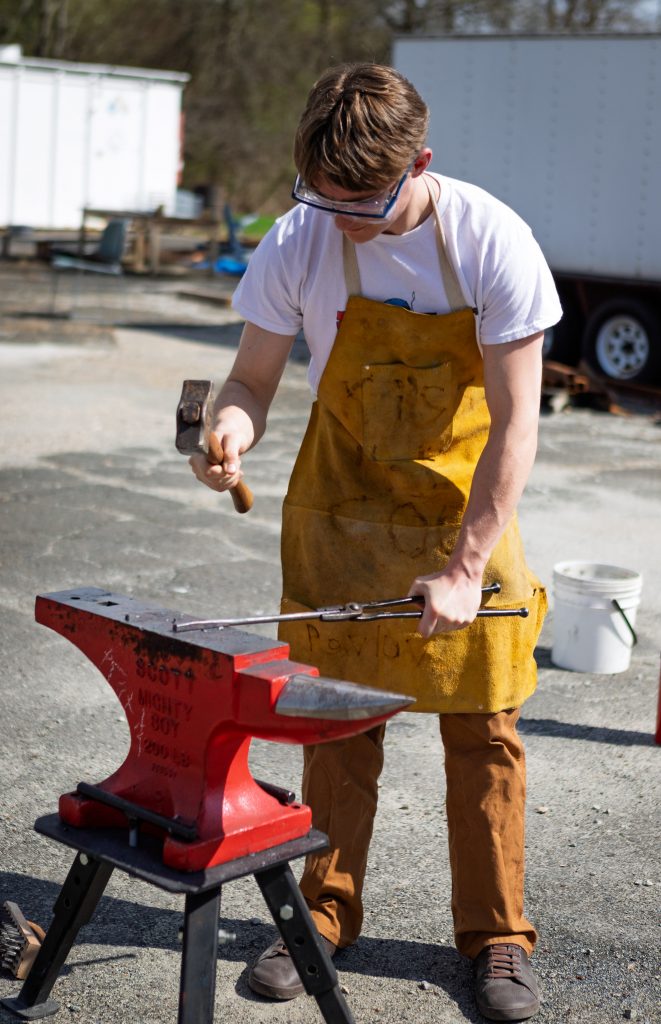 Student wearing safety goggles and apron hammers heated metal on an anvil during an outdoor metalworking session.