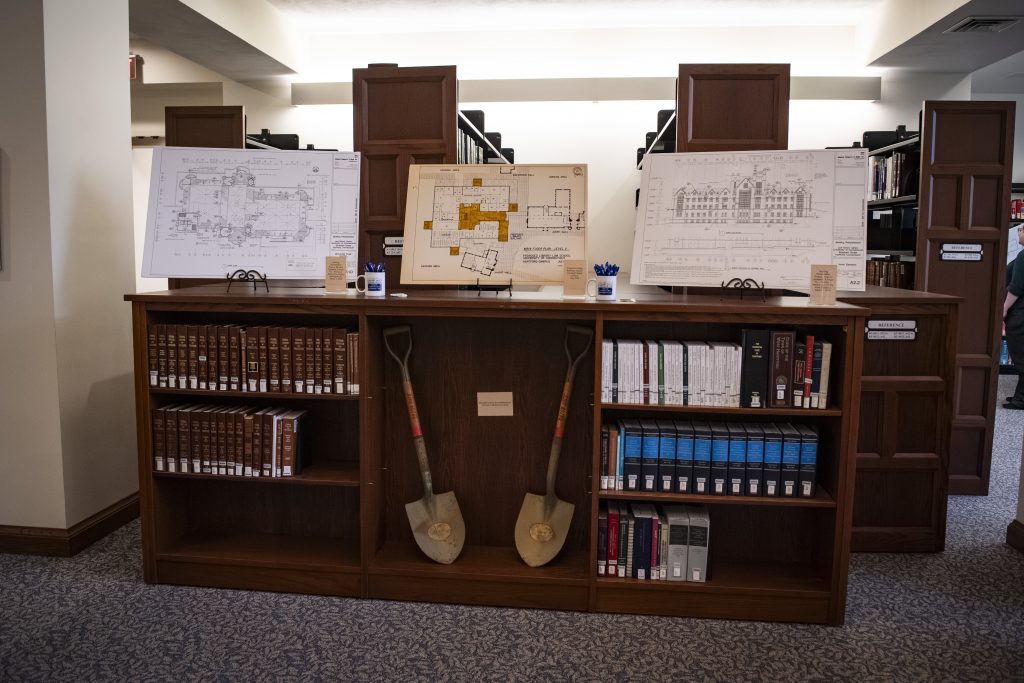 Two shovels are in the center of a three-section bookcase, with three sets of building plans propped upright on the top of the case.