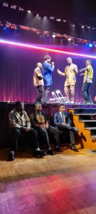 Three men sit in front of a stage while other men on stage model fashions.