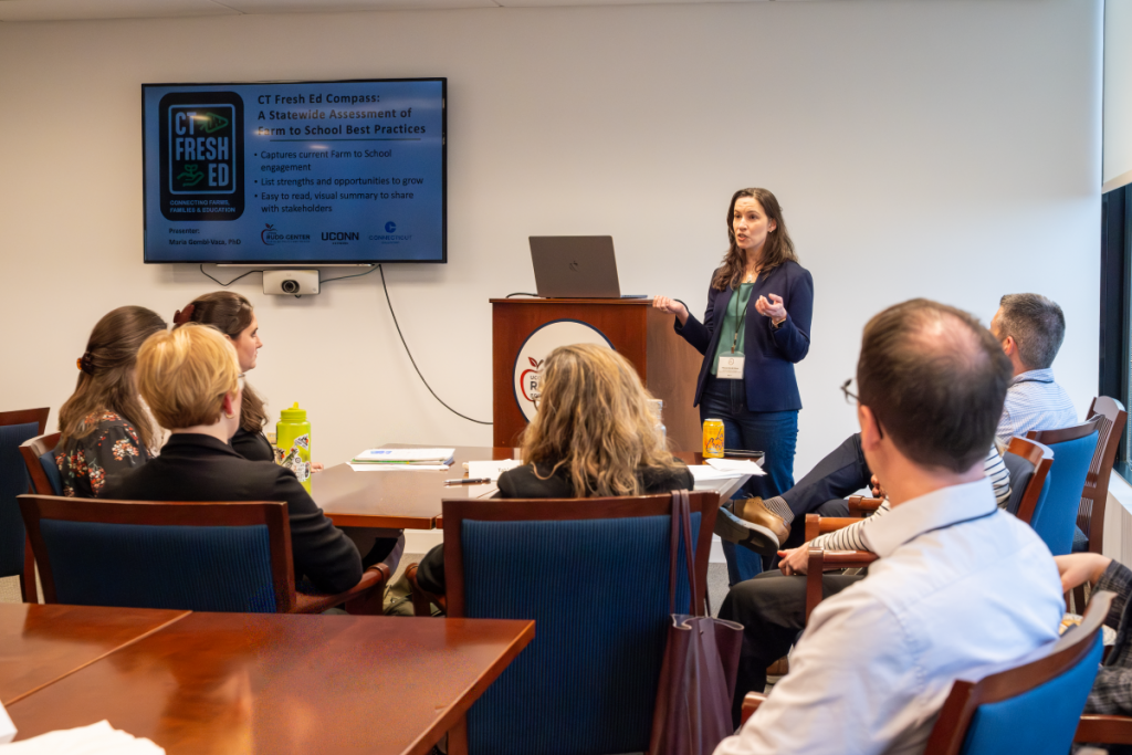 A woman stands before a video screen, addressing a room.