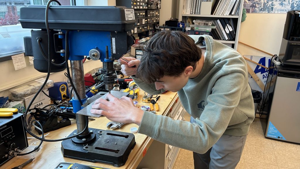 Student using a drill press to carefully machine a small metal component in a workshop.