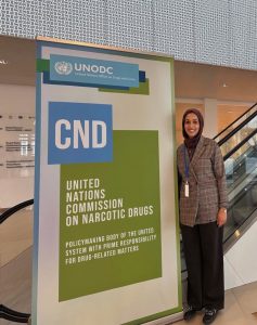A young woman stands next to a large sign showing she is at a meeting of the UN Commission on Narcotic Drugs.