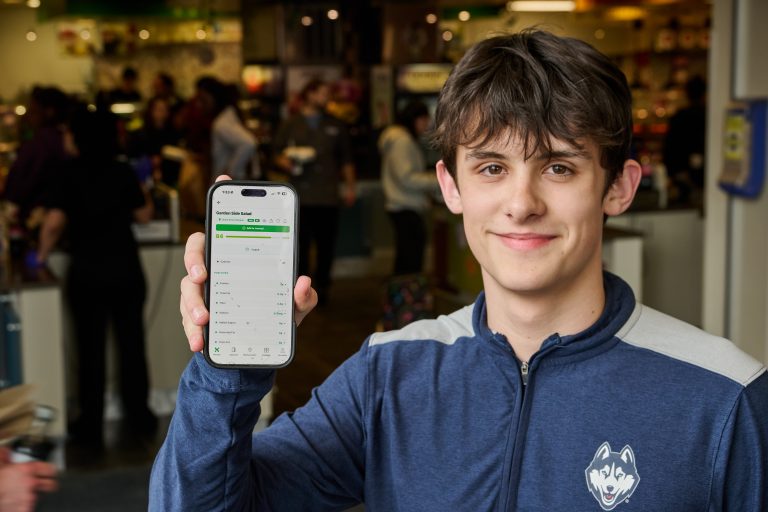 A young man with brown hair holds up a a phone to show an app on the screen