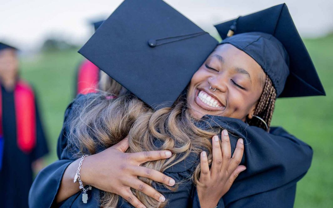 Two female graduates are seen hugging each other as they celebrate finally being done their education. They are dressed in formal black gowns and have caps on.