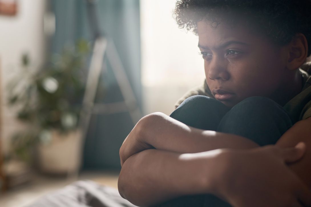 A child sits and hugs his knees to his chest. He has a tear in his eye