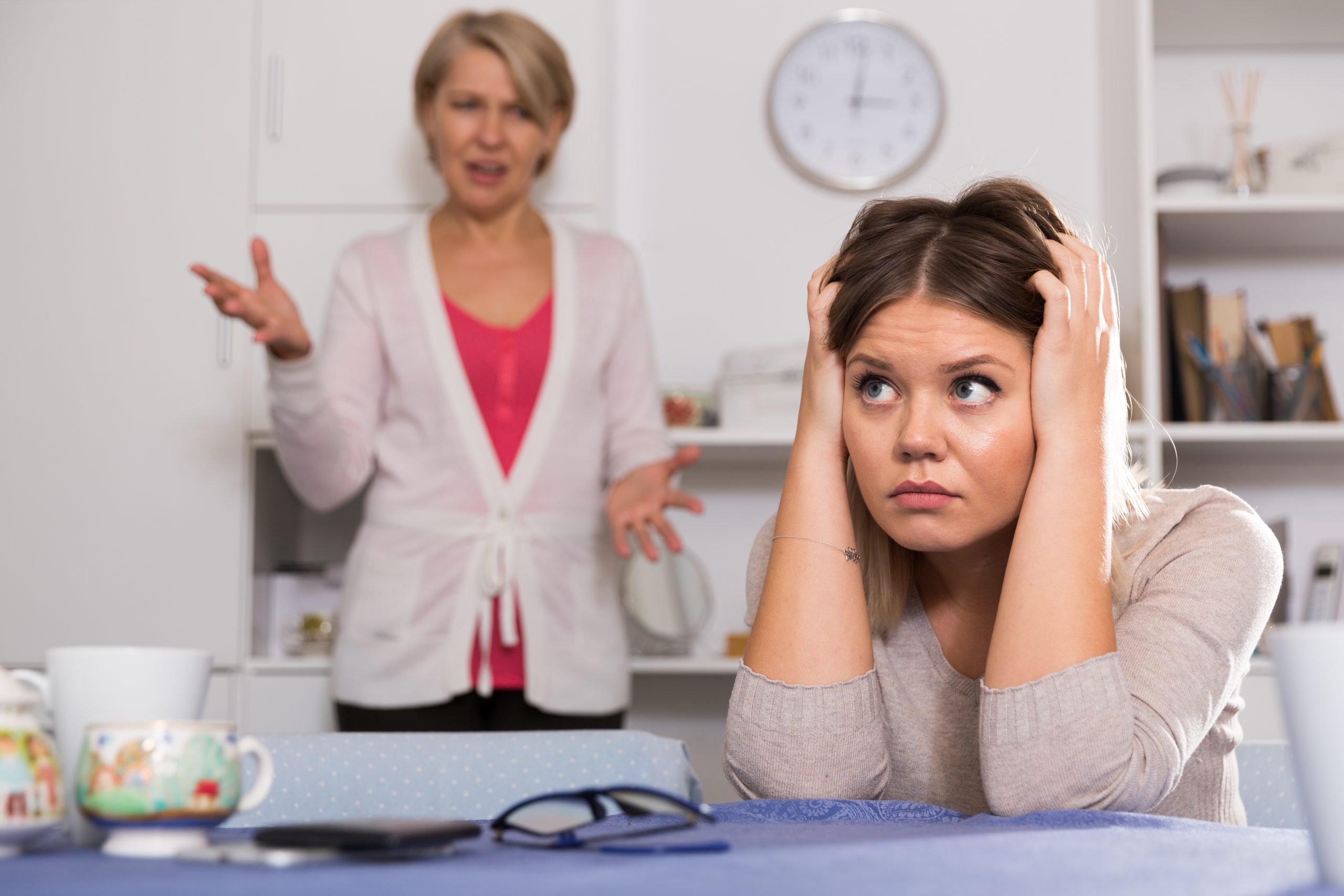 A teenager sits at a kitchen table holding her head in her hands while her mother teases her