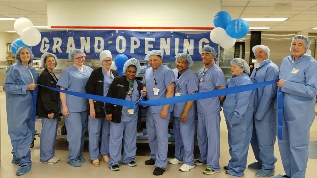 11 people in personal protective equipment gather for ribbon cutting in front of a 