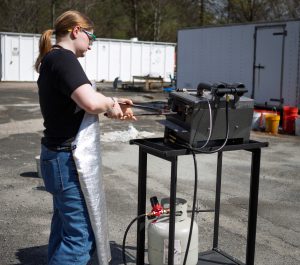A woman in protective gear works with forging equipment.
