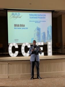 A studetn speaks in front of a stage and powerpoint to a group of people