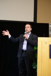 A man holding a microphone speaks on a stage next to a podium.