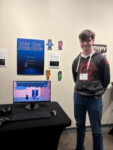 A boy poses in front of a display of his video game on a computer. 