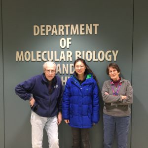 A man and two women stand in front of a wall with the words Department of Molecular Biology mounted on it.
