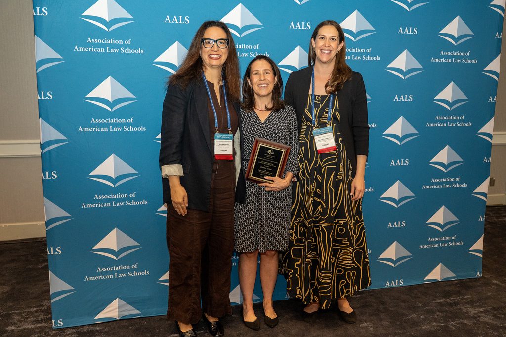 Three women standing in a line. The woman in the center is holding a plaque.