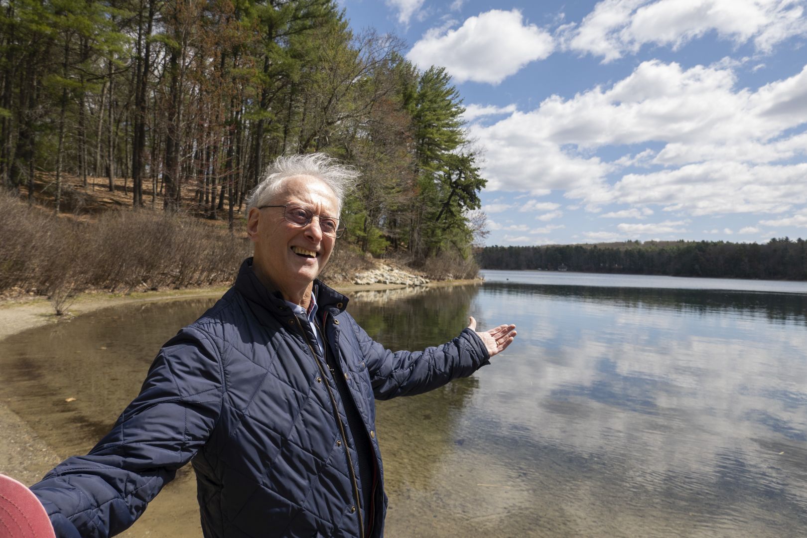 A person standing at the shore of a pond, with trees and a blue sky in the background.