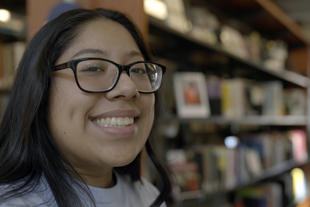 A woman with glasses smiles at the camera with books in the background.