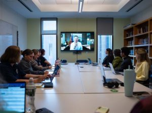 A group of people sitting on either side of a long table, with a person calling in on the video screen in front of them.