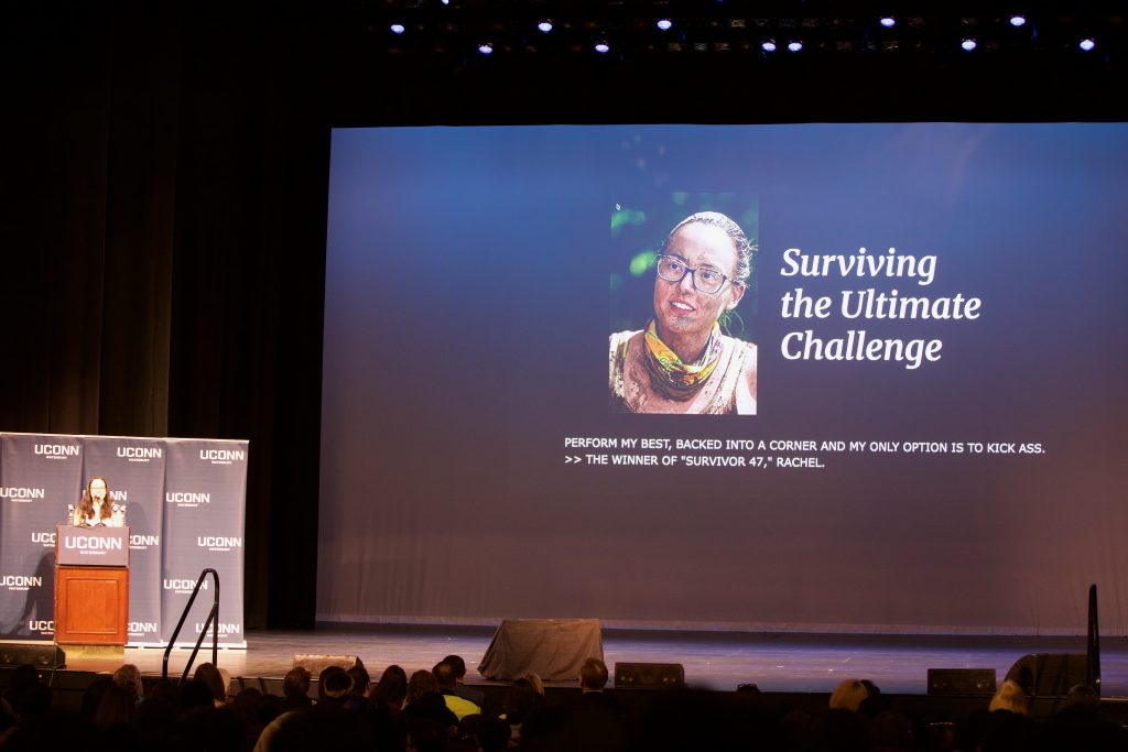 A woman speaks from a podium next to a large screen with her picture and the words "Surviving the Ultimate Challenge."
