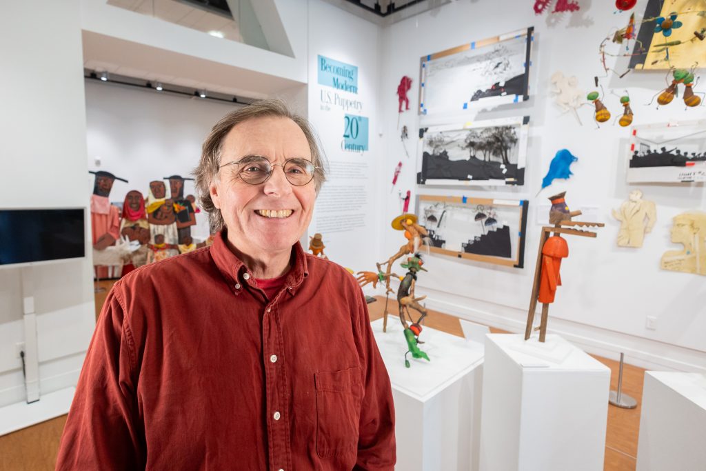 A man in a red dress shirt poses for a photo in front of a puppet display