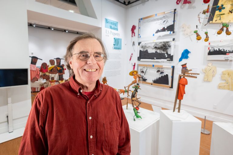 A man in a red dress shirt poses for a photo in front of a puppet display