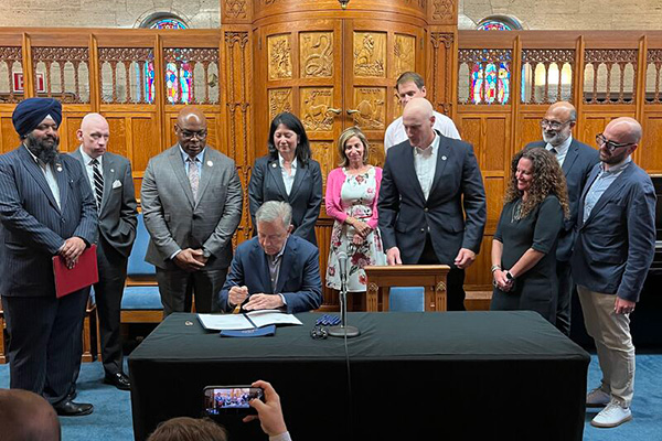 Gov. Ned Lamont signs a bill at a desk while supporters and officials look on behind him.