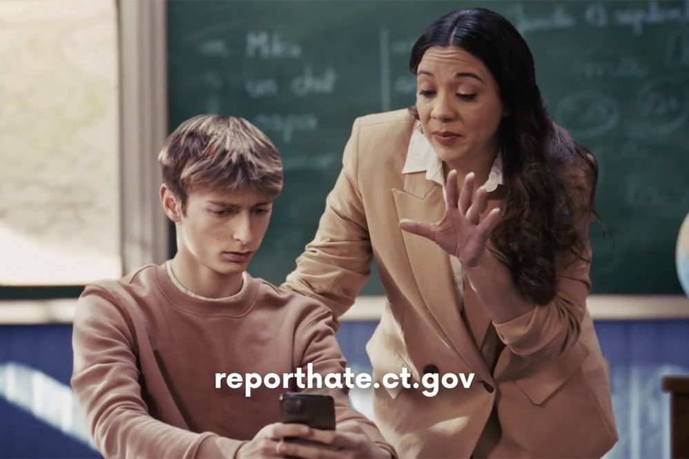 Instructor speaks with a student holding a cellphone beneath text reading “reporthate.ct.gov.”