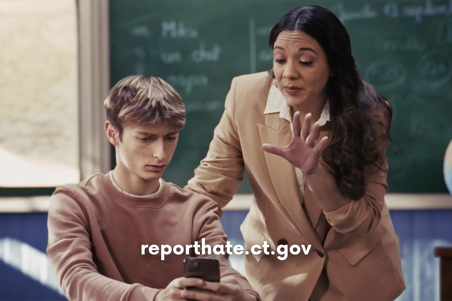Instructor speaks with a student holding a cellphone beneath text reading “reporthate.ct.gov.”