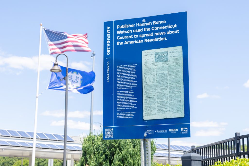 A Connecticut America 250 sign with information about the Hartford Courant with a Connecticut flag and U.S.A flag next to it
