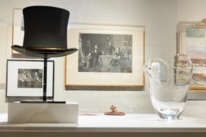 A portrait of the Lincoln family hangs behind a lead crystal Lincoln-themed vase, a top hat and a pile of American pennies