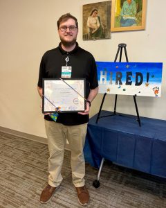 portrait man holding certificate next to sign that says "Hired!"