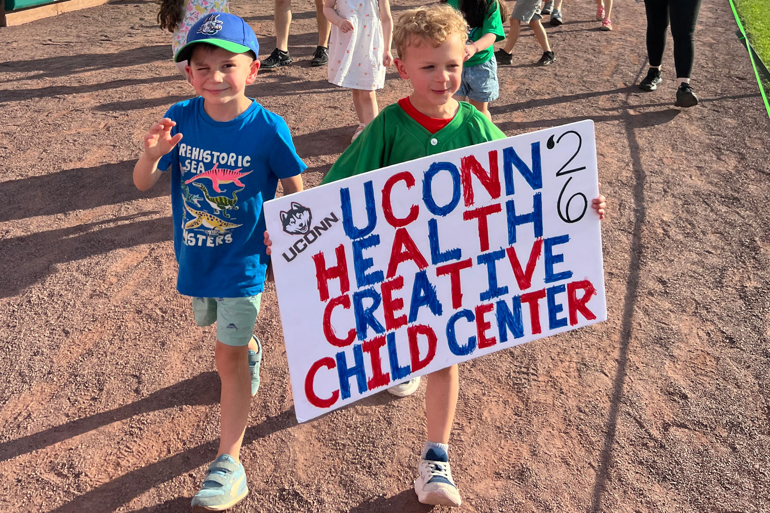 Young boys on baseball field carrying banner "UConn Health Creative Child Center ’26