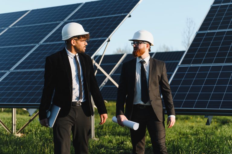 Two businessmen wearing suits and hard hats walking and discussing next to solar panels in a large solar farm.