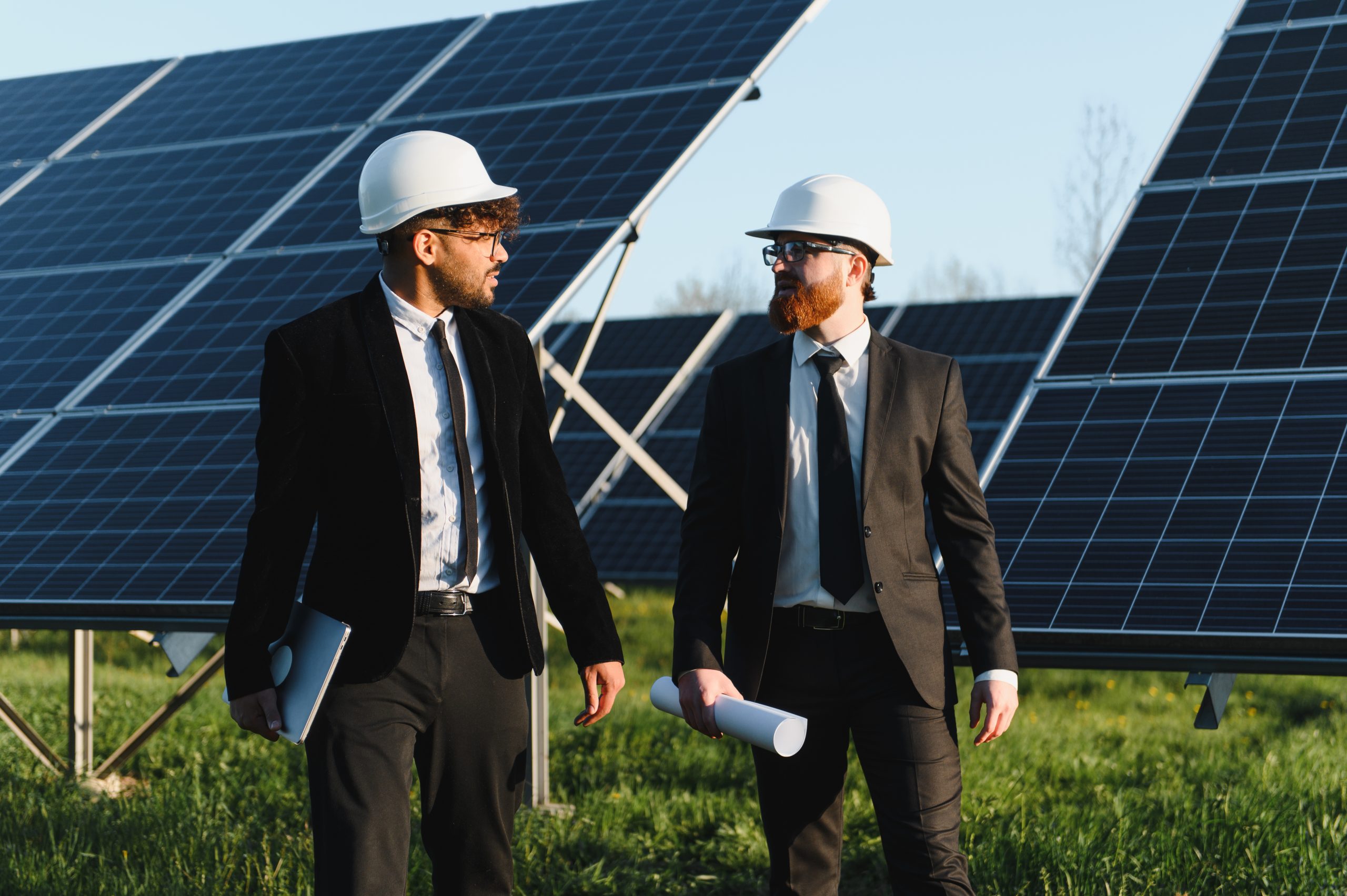 Two businessmen wearing suits and hard hats walking and discussing next to solar panels in a large solar farm.