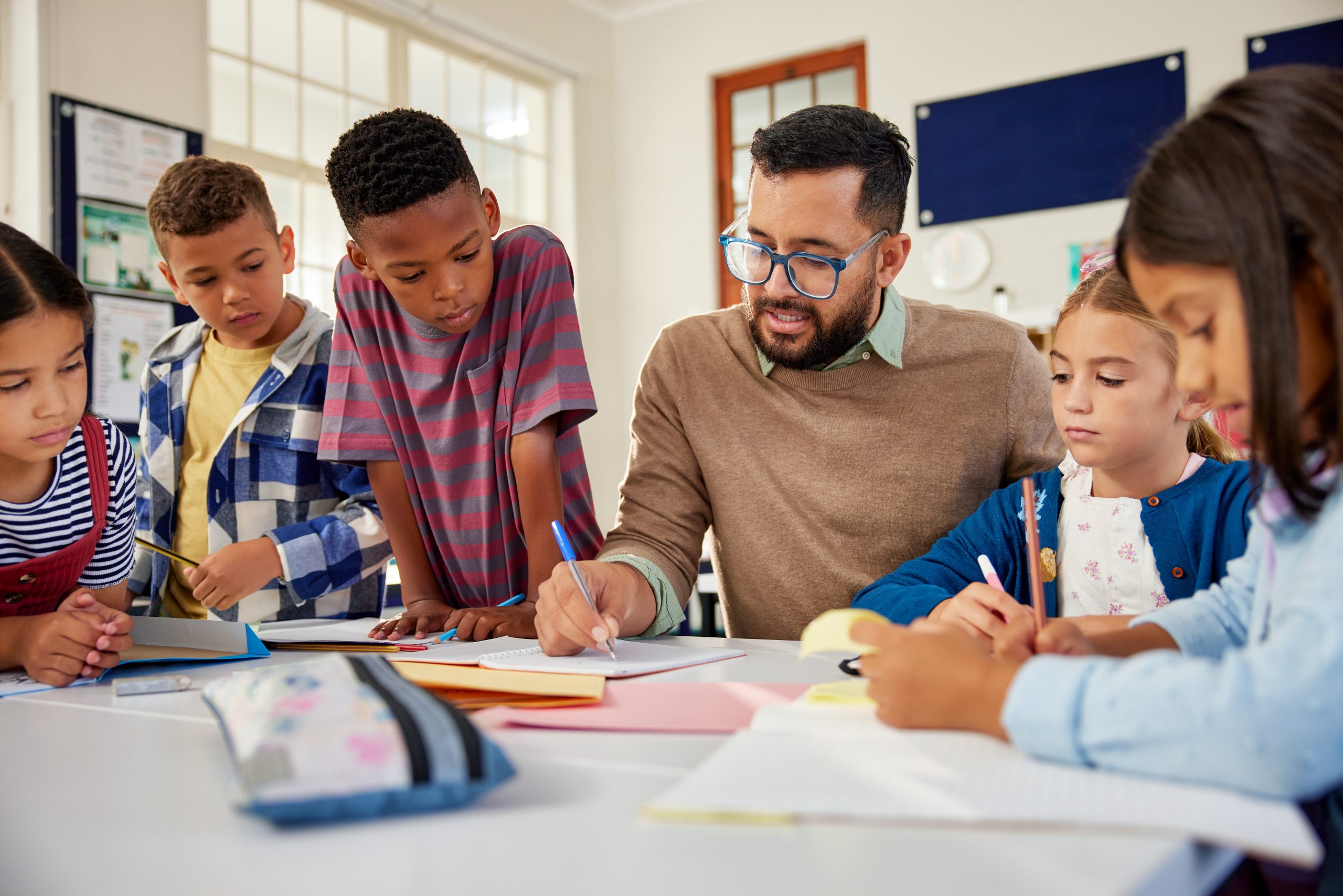 Hispanic male teacher wearing spectacles and checking student notebooks after test while talking to them in classroom.