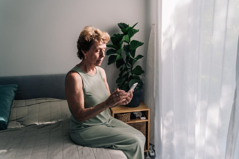 Elderly woman looking at her smartphone while sitting on a bed by a sunny window, surrounded by soft decor and a green plant in a pot.
